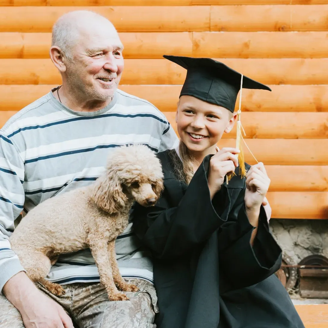 Man and a boy wearing graduation robes and hats, sitting on a bench with a dog between them