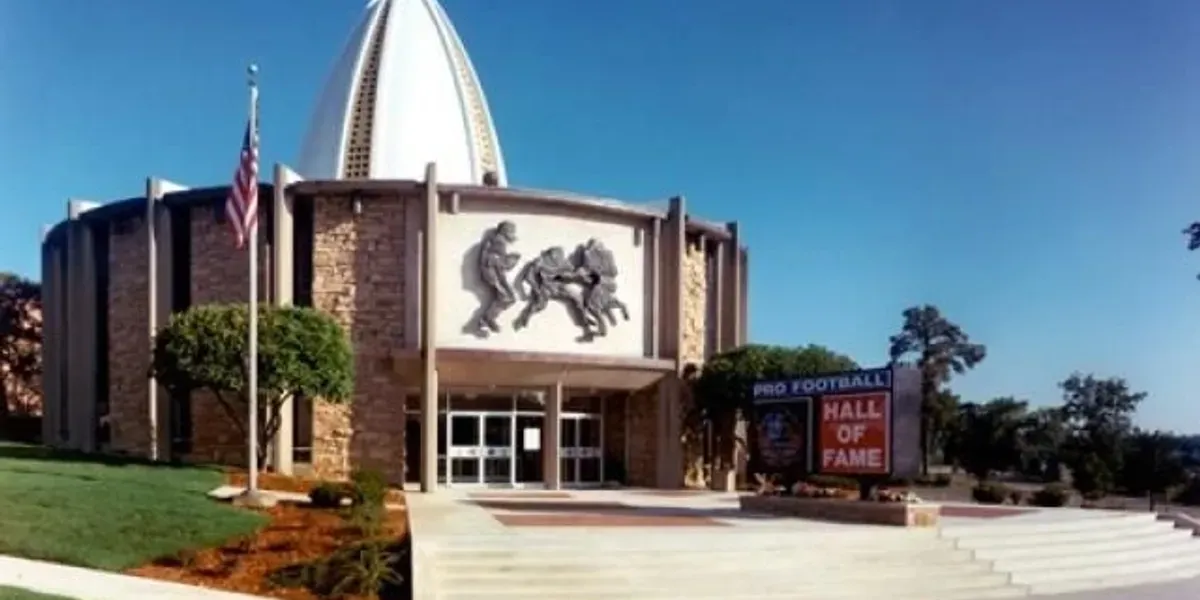 Large building with a flag on top of it, surrounded by trees, plants, grass, and a board with text