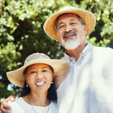 Older couple at the camera, both wearing hats and standing in front of a backdrop of trees