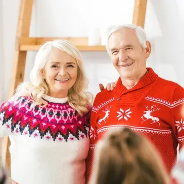 Older couple wearing Christmas sweaters, and standing in front of a group of people