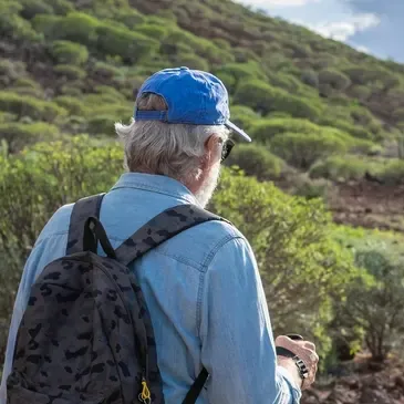 Man wearing a hat and a backpack, standing on a hill with trees in the background