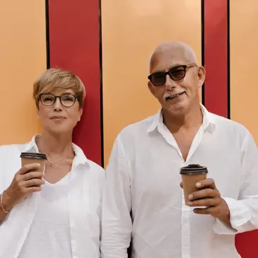Man and woman standing next to each other, both wearing white dresses and glasses, holding coffee cups in their hands