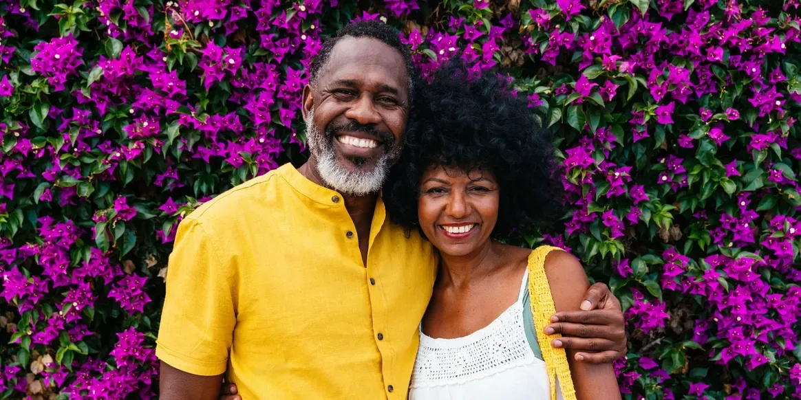 Man and woman standing next to each other in front of a wall of purple flowers