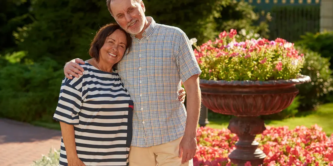 Man and woman standing next to each other in front of a fountain surrounded by lush greenery, including trees, plants