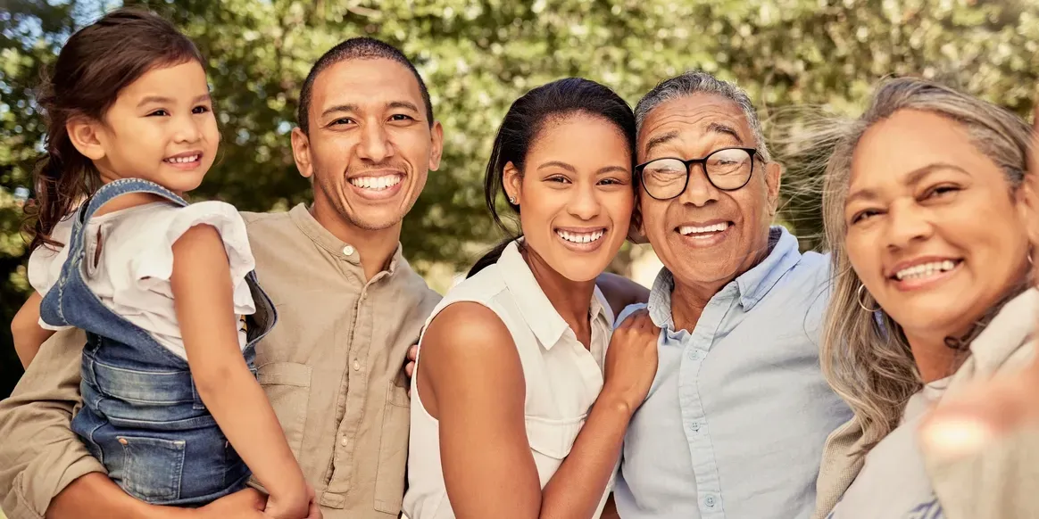 Family of four and posing for a photo in the park, with a man carrying a girl on his shoulders