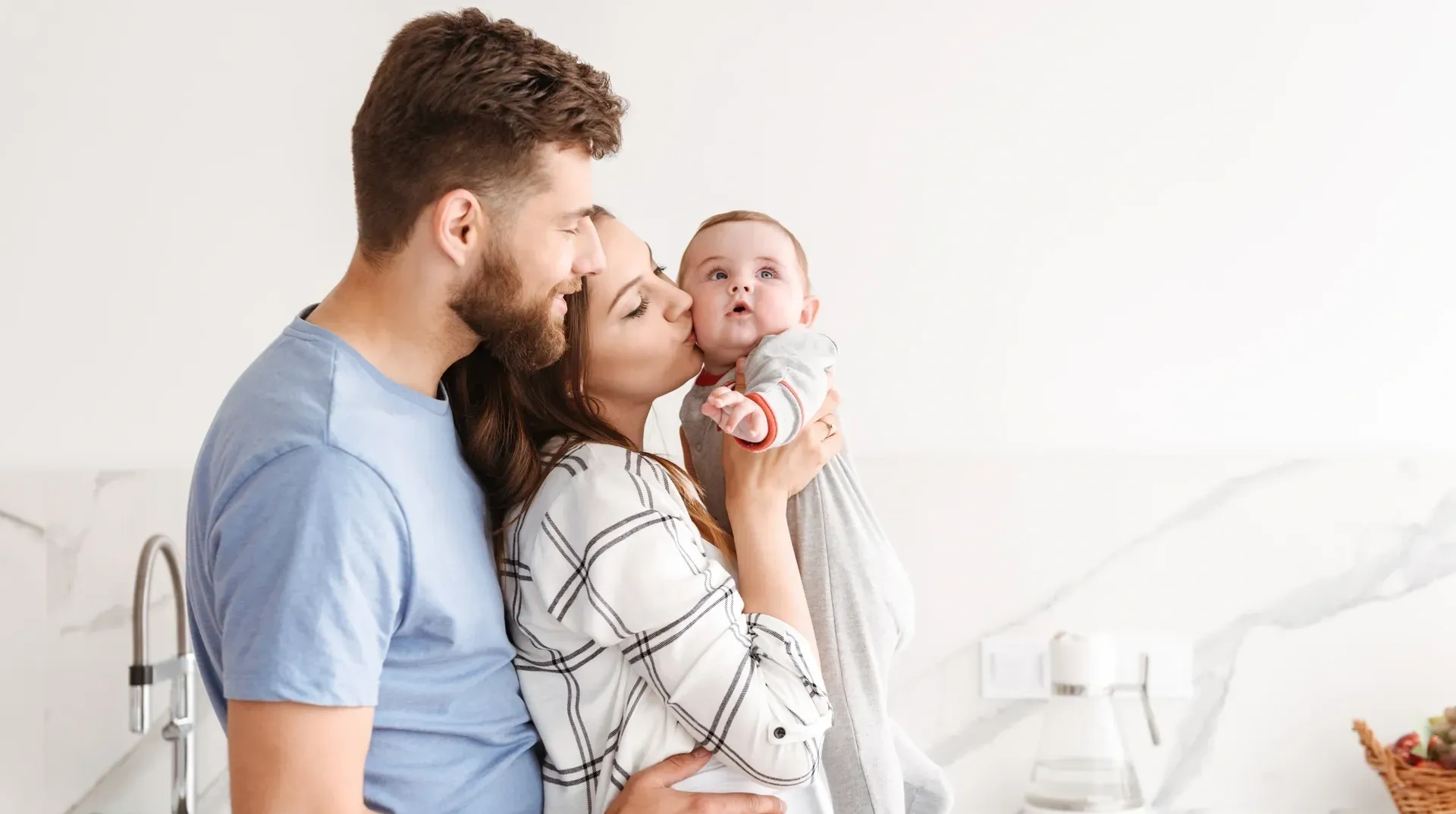 Man and woman holding a baby in a kitchen, with a basket of fruits on the right side